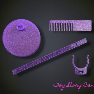 May include: A purple glittery doll stand set including a circular base with a central peg, a long vertical support rod, a U-shaped waist clip, and a small rectangular hair comb. The text Joy Story Canada is printed in pink at the bottom right.
