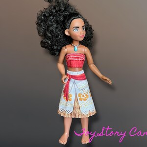 May include: A doll with dark curly hair, wearing a red and white top, a white skirt with orange floral patterns, and a turquoise necklace. The doll has a tan complexion and is posed against a brown gradient background.