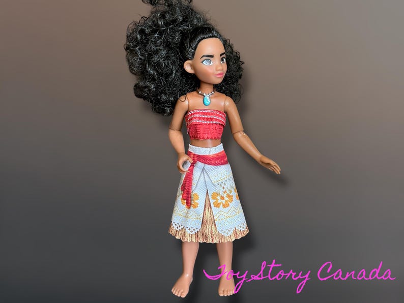 May include: A doll with dark curly hair, wearing a red and white top, a white skirt with orange floral patterns, and a turquoise necklace. The doll has a tan complexion and is posed against a brown gradient background.