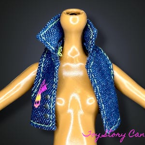 Peut inclure: Un gilet en jean bleu avec des coutures vert néon et un motif rose et jaune. Le gilet est présenté sur un torse de poupée. Le gilet a trois boutons noirs et un col. Le fond est gris foncé.