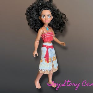 May include: A doll of a character with dark curly hair, wearing a red top and a white skirt with a red sash. The doll has a turquoise necklace and is standing on a brown surface.