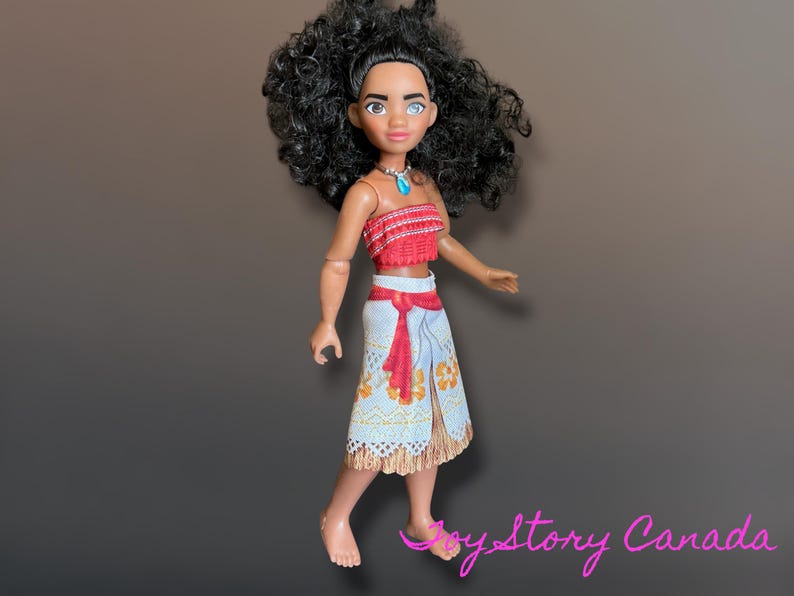 May include: A doll of a character with dark curly hair, wearing a red top and a white skirt with a red sash. The doll has a turquoise necklace and is standing on a brown surface.
