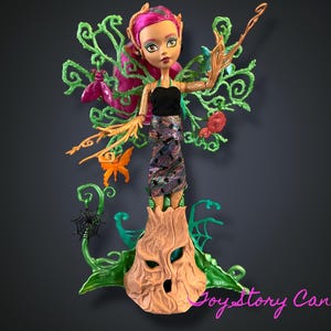 Pode incluir: Uma boneca Monster High verde e marrom com cabelo rosa e um vestido preto e verde. A boneca está em pé em um tronco de árvore com videiras e folhas. A boneca tem uma borboleta no ombro e uma teia de aranha no tronco da árvore.