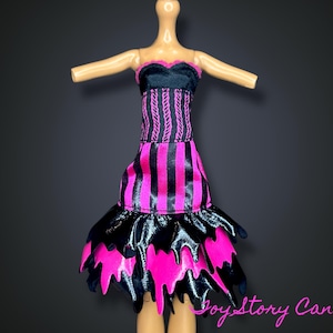 Puede incluir: Un vestido de muñeca con un corpiño negro y una falda a rayas rosas y negras. La falda tiene un dobladillo en capas y dentado. El vestido está diseñado con un top sin tirantes y está hecho de tela brillante. El vestido es un diseño vibrante y llamativo.