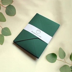 May include: A dark green envelope secured with a white paper band featuring a circular logo and the text "Thanks for your order" in black script. The envelope is positioned diagonally, showcasing its pointed flap and smooth matte texture.