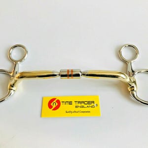 May include: A horse bit with a gold and silver finish. The bit has a central mouthpiece with orange stripes and two circular rings on either side. A yellow tag with the text "TIME TRADER ENGLAND" is below the bit.