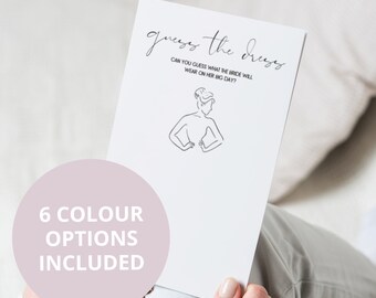 May include: A white card with the text "guess the dress" and a line drawing of a woman in a dress. The text below the drawing says "Can you guess what the bride will wear on her big day?"  The card also has the text "6 colour options included" in a pink circle.