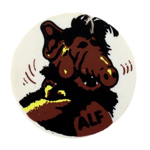 May include: A white round sticker with a cartoon image of ALF, an alien from the TV show ALF. ALF is brown and black with a large nose and big eyes. The sticker has the word "ALF" written in brown letters on the bottom right corner.