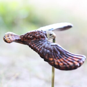 以下が含まれることがあります： 飛行中の鷹の木彫りの詳細。鳥は茶色とタン色の羽を持ち、頭とくちばしは明るい色です。翼は大きく広げられ、彫刻は細い金色の棒に取り付けられています。