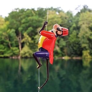 May include: A figurine of a person climbing a rope, with a red coat, yellow shirt, and blue trousers. The head is covered with a red hat. The background shows a lake and green trees.