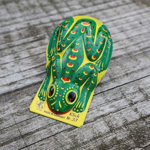 Könnte beinhalten: Ein Vintage-Spielzeugfrosch mit grünem Körper, gelbem Kopf und roten Punkten. Der Frosch hat schwarze Augen und ist mit "Klick Nr.33 Made in Germany" beschriftet. Das Spielzeug ist aus Metall und liegt auf einer Holzoberfläche.