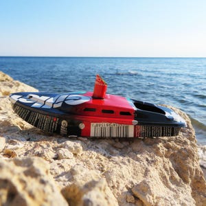 Può includere: Una barca giocattolo realizzata con una lattina di metallo riciclata, dipinta in rosso e nero con dettagli bianchi. La barca è posizionata su una roccia, con il mare e un cielo blu sullo sfondo. La lunghezza della barca è di circa 15 cm.