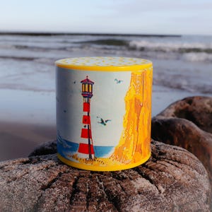 Puede incluir: Un recipiente cilíndrico con tapa y base amarillas. El recipiente presenta un diseño de faro con rayas rojas y blancas, un mar azul y acantilados amarillos. El fondo muestra una playa y el océano.