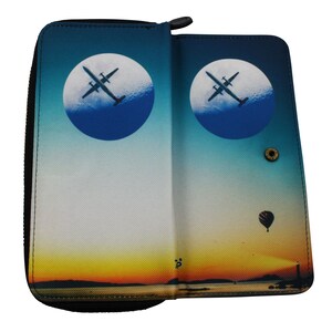 May include: A black zippered wallet with a blue, white and yellow sunset scene. The wallet has two circular images of aeroplanes flying in front of a blue circle. A hot air balloon is flying in the sky above the sunset.