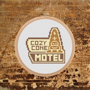 May include: A cross-stitch embroidery in a wooden hoop features the words "COZY CONE MOTEL" in a pixelated design. The design includes a stylized motel sign with a cone-shaped structure in shades of brown and beige, set against a white background.