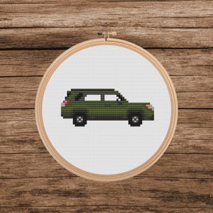 May include: A cross-stitch embroidery hoop featuring a pixelated green SUV design. The vehicle has black wheels and a red taillight. The embroidery is set against a white background, framed by a wooden hoop, and placed on a wooden surface.