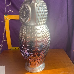 May include: A silver and iridescent glass owl figurine with a textured surface. The owl has a round head with a large eye and a detailed beak. The body is covered in small bumps and has a feathered design.