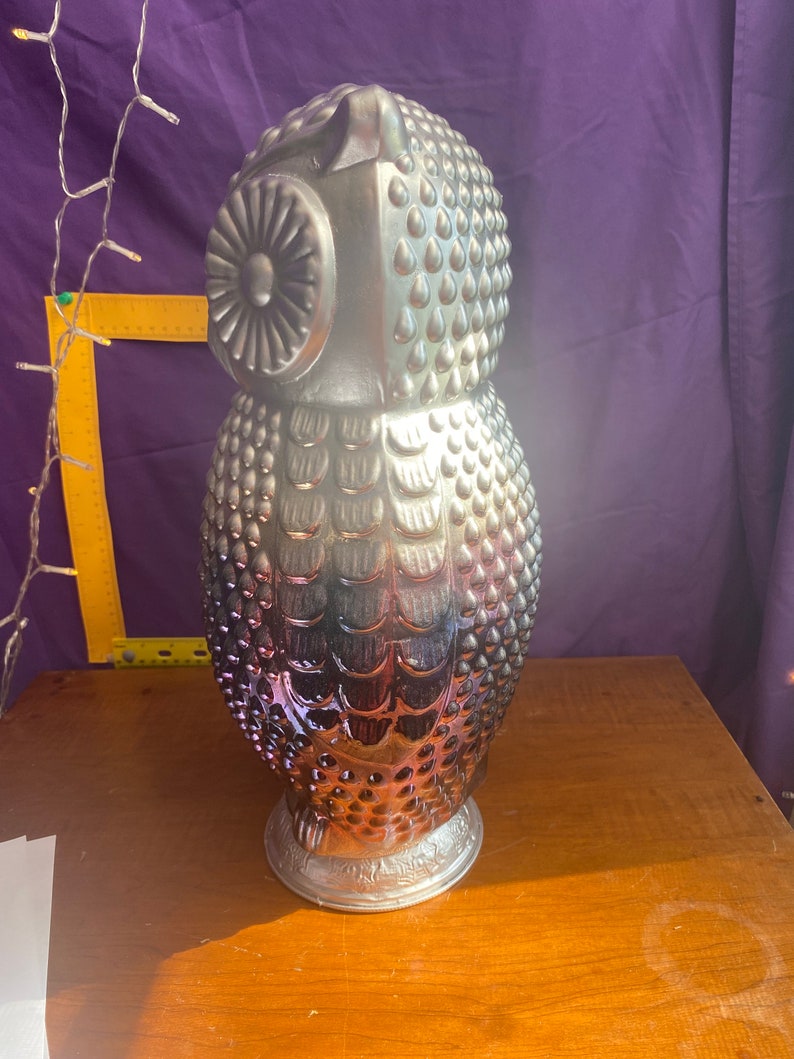 May include: A silver and iridescent glass owl figurine with a textured surface. The owl has a round head with a large eye and a detailed beak. The body is covered in small bumps and has a feathered design.