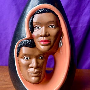 May include: A black ceramic vase with a smooth finish featuring a large, oval-shaped cutout in the center. Inside the cutout are two sculpted, three-dimensional faces of individuals with brown skin tones, pink lips, and dark hair, set against a peach-colored border.