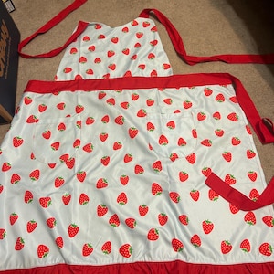 May include: A light blue apron featuring a repeating pattern of red strawberries with green leaves. The apron includes a solid red waistband, two front pockets, red fabric ties at the neck and waist, and a ruffled red hem along the bottom edge.