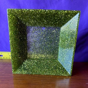 May include: A square green glitter picture frame with a clear glass insert. The frame is made of resin and has a sparkly finish.