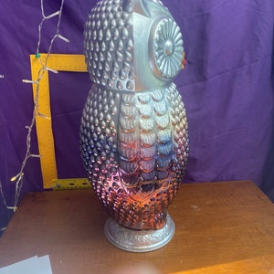 May include: A silver and iridescent owl figurine with a textured surface. The owl has a round head with a large eye and a small beak. The owl is standing on a base with a decorative design.