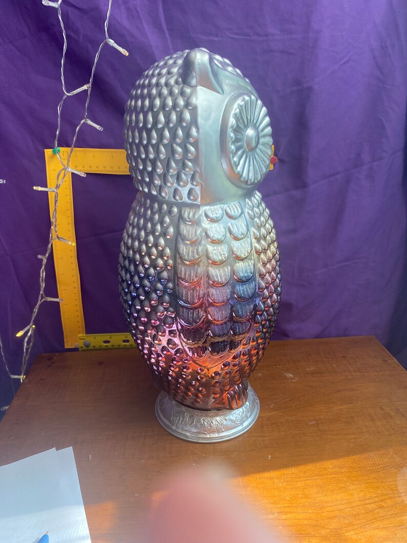 May include: A silver and iridescent owl figurine with a textured surface. The owl has a round head with a large eye and a small beak. The owl is standing on a base with a decorative design.
