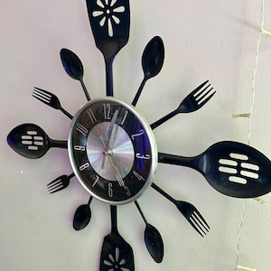 May include: A black and silver kitchen clock with a unique design. The clock face is surrounded by black utensils, including spoons, forks, and spatulas. The clock hands are silver, and the numbers are black.
