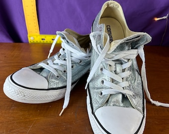 May include: A pair of silver glitter Converse Chuck Taylor All Star low-top sneakers with white laces and a white rubber toe cap. The shoes are on a wooden surface with a purple background.