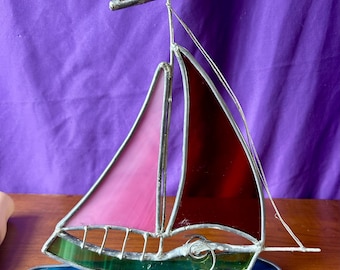May include: Stained glass sailboat with a red pennant, pink and red sails, and a blue and green base. The sailboat is made with lead came and sits on a wooden surface against a purple background. The sails are translucent.