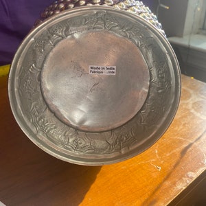 May include: A silver metal vase with a textured, embossed design. The vase has a round base and a wide opening. The bottom of the vase has a label that reads "Made in India Fabrique - Inde".