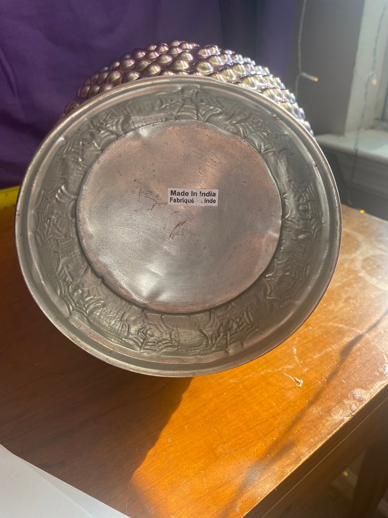 May include: A silver metal vase with a textured, embossed design. The vase has a round base and a wide opening. The bottom of the vase has a label that reads "Made in India Fabrique - Inde".