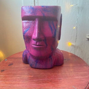 May include: A pink and blue ceramic planter shaped like a human face. The planter has a marbled design and is sitting on a wooden table.