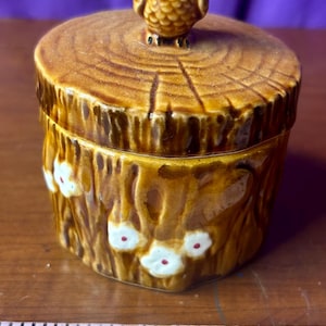 May include: A ceramic owl-topped trinket box. The box is brown with a tree stump design and white flower accents. The owl has yellow eyes and is perched on the lid. The box measures approximately 4 inches in height.