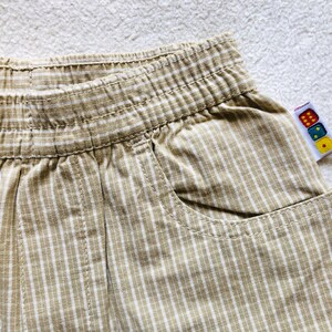 K&ouml;nnte beinhalten: Beige und wei&szlig;e gestreifte Baumwollhose mit einer einzigen Tasche auf der rechten Seite. Die Hose hat einen elastischen Bund und ein Etikett mit bunten W&uuml;rfeln.
