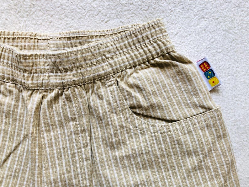 K&ouml;nnte beinhalten: Beige und wei&szlig;e gestreifte Baumwollhose mit einer einzigen Tasche auf der rechten Seite. Die Hose hat einen elastischen Bund und ein Etikett mit bunten W&uuml;rfeln.