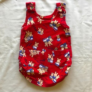 May include: Red corduroy baby onesie with a pattern of cartoon bunnies. The onesie has two pockets and a sleeveless design.