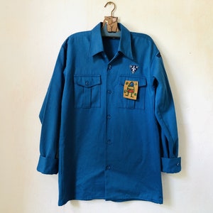 Op de afbeelding: Een blauw overhemd met lange mouwen en een patch aan de rechterkant. De patch heeft een symbool en een tekening van een persoon.