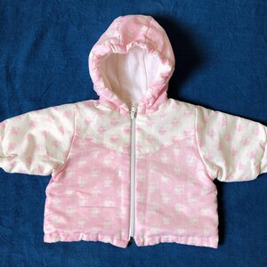 Puede incluir: Chaqueta con capucha rosa y blanca para bebé. La chaqueta tiene una cremallera completa y un patrón de pequeñas formas repetidas. La capucha y el forro son blancos. La chaqueta es de material suave.