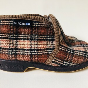 May include: Brown and black plaid slipper with a black rubber sole. The slipper has a brown cuff and the word "SUOMI" printed on the side.