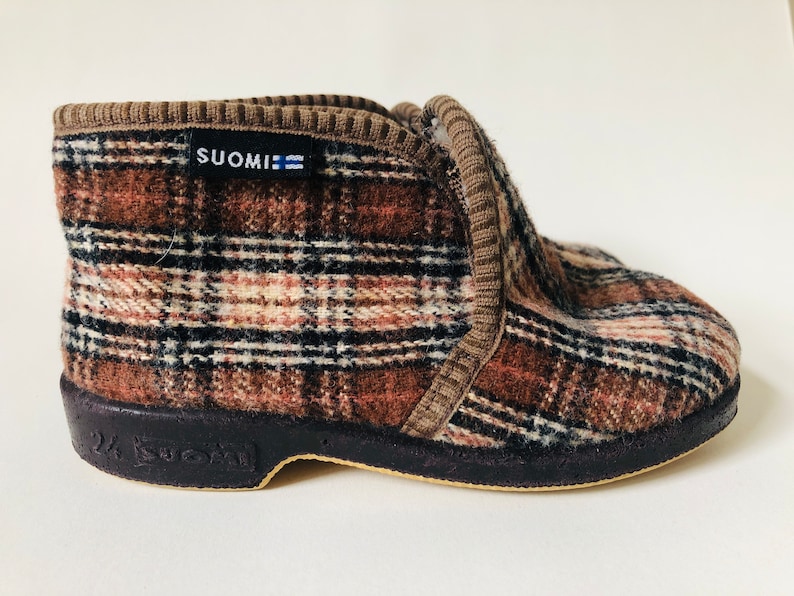 May include: Brown and black plaid slipper with a black rubber sole. The slipper has a brown cuff and the word "SUOMI" printed on the side.