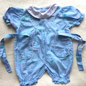 May include: A light blue baby onesie with a white collar and short sleeves. The onesie has a tie at the waist and features a colorful print of abstract shapes and dolphins.