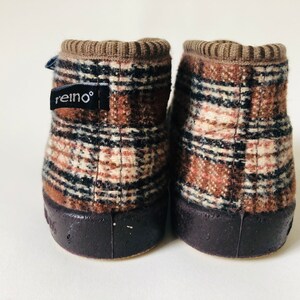 May include: A pair of brown and beige plaid slipper boots with black trim. The boots have a brown rubber sole and a brown fabric cuff. The left boot has a black label with the word "reino" on it.