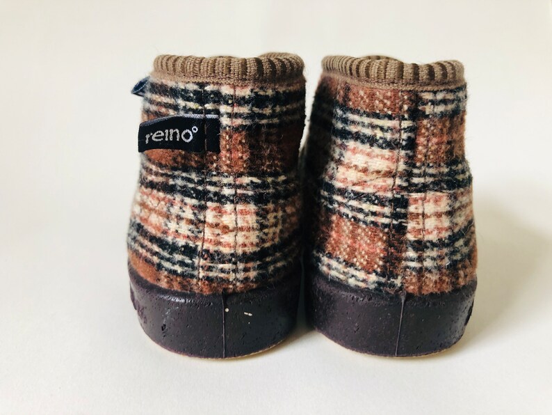May include: A pair of brown and beige plaid slipper boots with black trim. The boots have a brown rubber sole and a brown fabric cuff. The left boot has a black label with the word "reino" on it.
