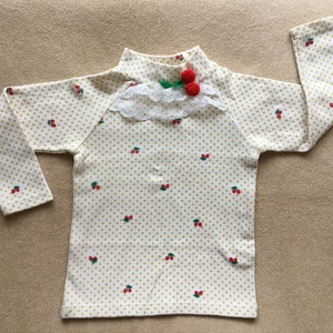 May include: A long-sleeved, cream-colored shirt with a pattern of small yellow dots and red cherries. The shirt has a high neckline, a white lace collar, and a pair of red cherry embellishments. The sleeves are long and the shirt is made of a soft fabric.