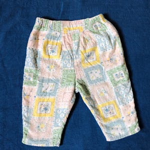Może przedstawiać: Spodnie dla niemowląt z elastyczną talią. Spodnie mają wzór patchworkowy z kwadratami w pastelowych kolorach: różowym, żółtym, zielonym i niebieskim. Każdy kwadrat ma kwiatowy lub wzorzysty wzór. Spodnie są wykonane z miękkiej tkaniny.