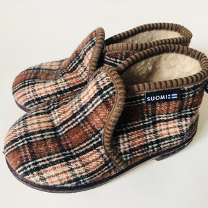 May include: Brown and beige plaid slippers with a brown fleece lining. The slippers have a brown trim and a blue label that says "SUOMI".