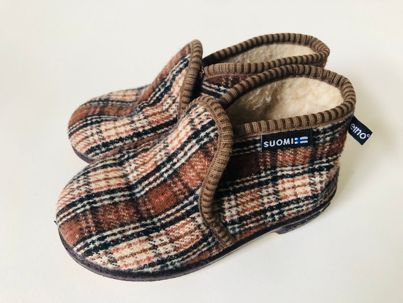 May include: Brown and beige plaid slippers with a brown fleece lining. The slippers have a brown trim and a blue label that says "SUOMI".