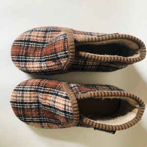 May include: A pair of brown and black plaid slippers with a brown fleece lining. The slippers have a brown elastic band at the top and the brand name "Simons" is visible on the bottom of one slipper.