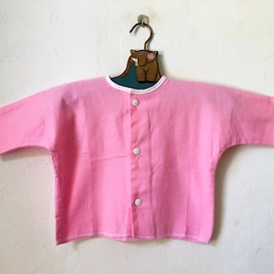 Peut inclure: Une chemise boutonnée rose avec une bordure blanche autour du col et des poignets. La chemise est suspendue à un cintre en bois avec un motif d'éléphant peint.